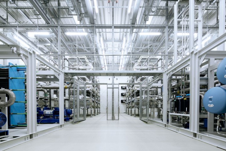 Innenansicht einer STATCOM-Anlage (Static Synchronous Compensator), die für die Regelung der Netzspannung eingesetzt wird. Die Halle ist hell beleuchtet und symmetrisch angeordnet. Sie ist geprägt von weißen Metallrahmen, Gitterstrukturen und technischer Infrastruktur wie Rohren, Ventilen und Maschinen. Die technische Ausrüstung ist in Reihen geordnet, und über der Anlage verlaufen Kabelkanäle sowie große LED-Leuchten, die den Raum gleichmäßig ausleuchten.