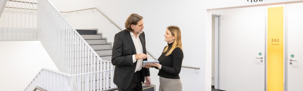 Gesprächssituation: Johanna Volk und Prof. Dr. Andreas Löschel stehen in Geschäftskleidung in einem hellen Treppenhaus. Frau Volk hält ein Tablet, Herr Lösch zeigt darauf. Beide schauen sich an.
